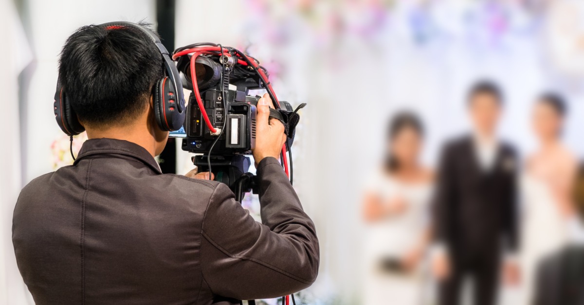 A man behind a video camera that's pointed at the bride and groom. The man is wearing a high-tech headset.
