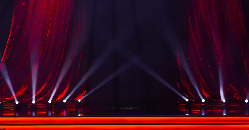 A red curtain that's hanging down on the surface of a stage. There are a bunch of spotlights sitting in front of them.
