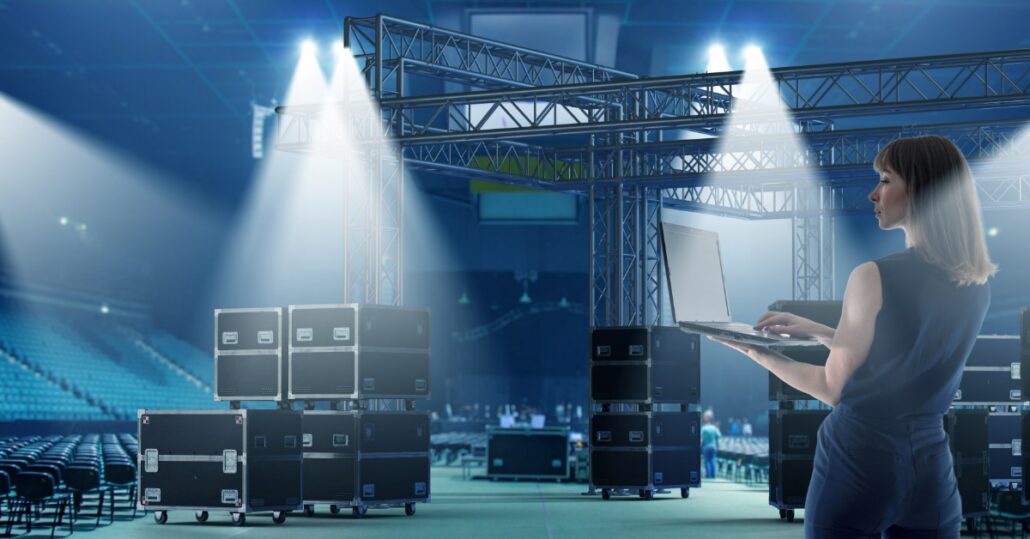 A stage coordinator uses a laptop near the lighting truss and equipment cases inside a concert venue during setup.
