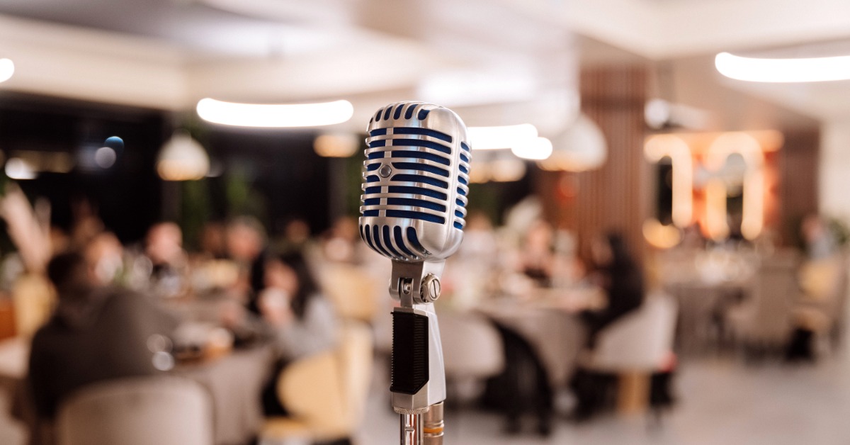 A retro microphone standing on a stage, ready for use. There are a bunch of people sitting at tables behind it.