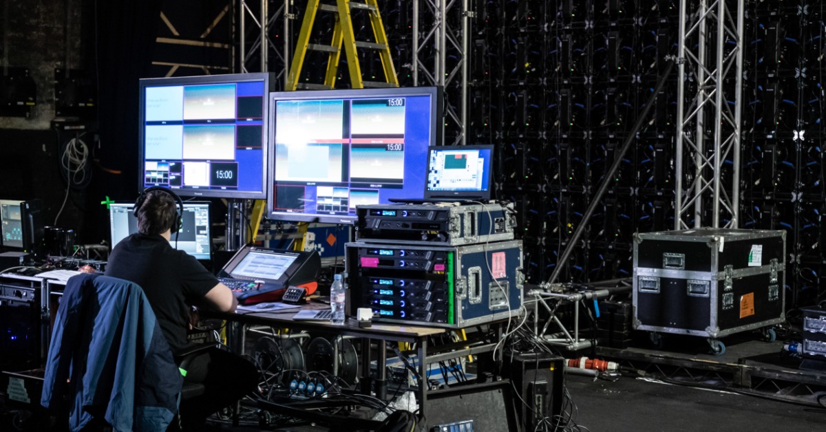 Backstage audio visual equipment is shown with an operator working with the screens, cables, and other control gear.