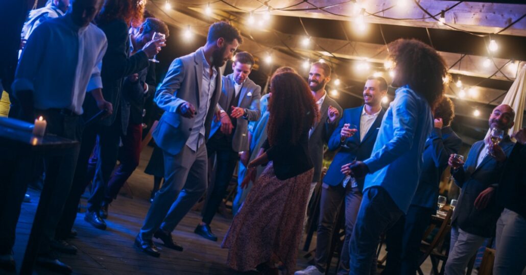 People dancing together at an evening wedding reception, with colorful lighting and tables arranged behind them.