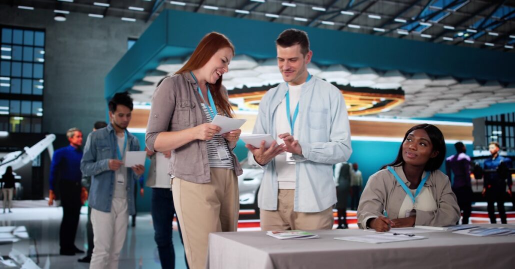 A saleswoman presenting materials to two other people at a trade show booth inside a large exhibition hall.