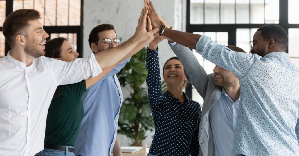 People standing in a circle with their hands high-fiving in the middle. They are smiling and wearing business casual clothes.