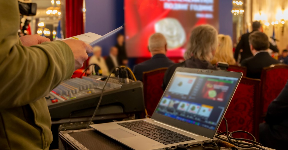 A laptop and audio mixer is on a table beside a person. Seated attendees and a large screen are in the background.