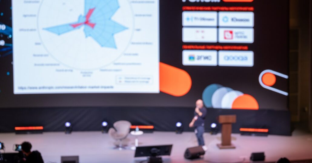 A speaker stands on a lit auditorium stage before large digital screens while an audience watches from rows of seats.