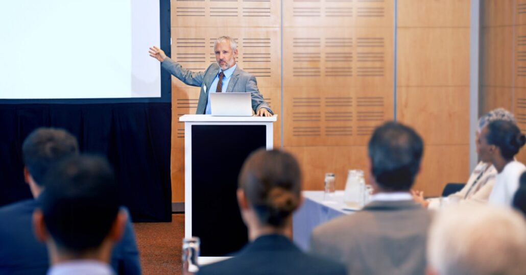 A man in a suit stands at a podium with a laptop. He's pointing toward a presentation screen in front of seated individuals.