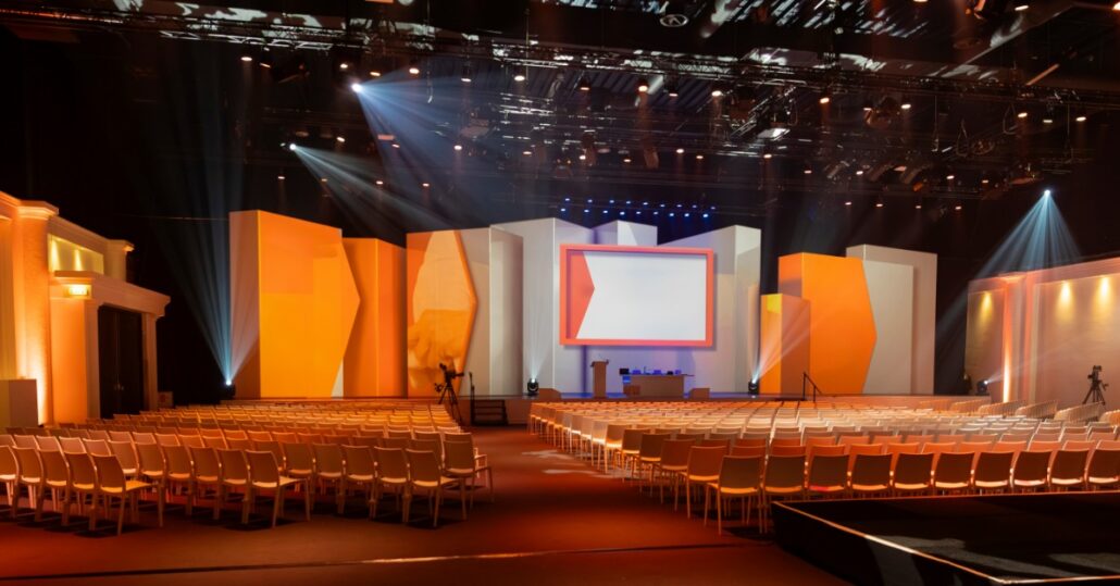 Rows of white chairs face a lit auditorium stage with orange panels, a central screen, a podium, and overhead spotlights.