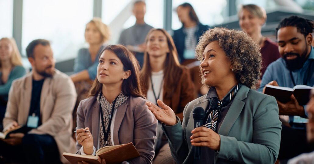 A woman holding a microphone gestures while another woman holds a pen and an open notebook among seated attendees.