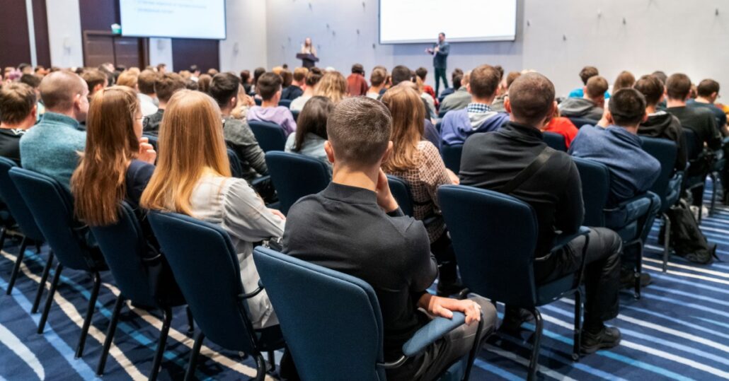 Rows of attendees sit in blue chairs facing two presenters and large projection screens in a conference room.
