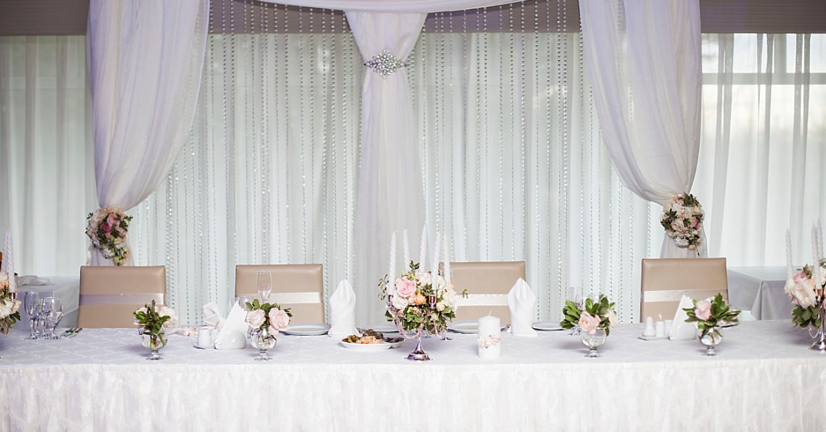 A long white table holds flowers, glasses, plates, napkins, and candles beneath draped fabric and hanging beads.